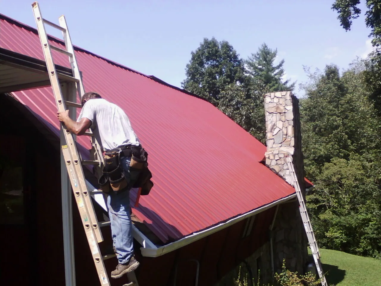 Roofer on a ladder beside a red metal roof and stone chimney — Soffit & Fascia Repair by Roofing Brothers in Wexford